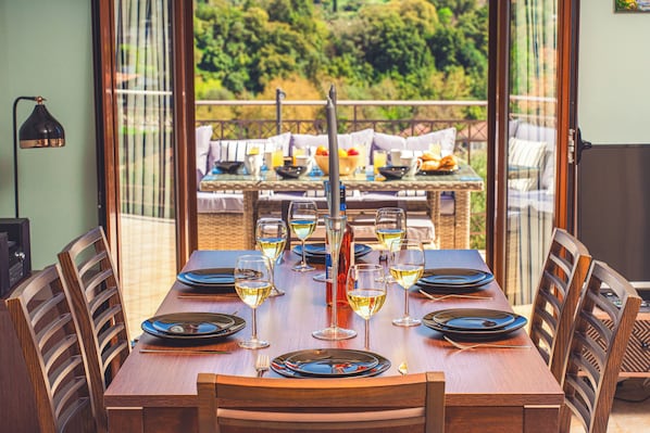 A dining table set for six with wine glasses, plates, and cutlery, overlooking a garden.