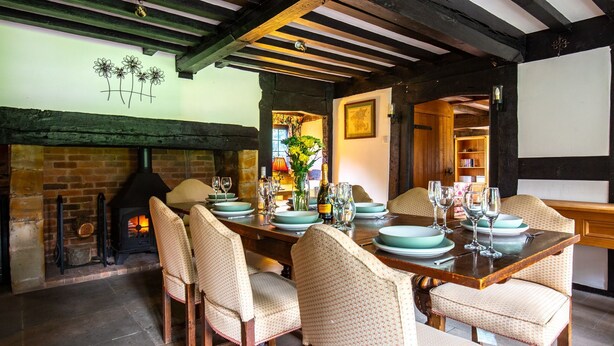 A dining area with a brick fireplace, wooden beams, and a table set for six with chairs.
