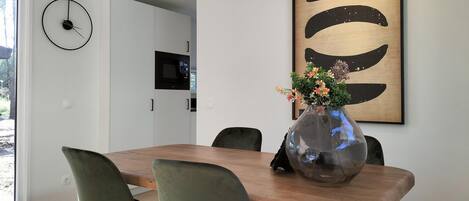 Un comedor moderno con una mesa de madera, sillas tapizadas en verde, un cuadro abstracto grande y un jarrón con flores.
