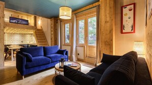 A cozy living room with a blue sofa, a black sofa, and a wooden dining table with chairs. There is a staircase leading to an upper level, and a large window with curtains. - Loft Bellevue Remonté | Centre | Piscine | Parking (Les Houches)