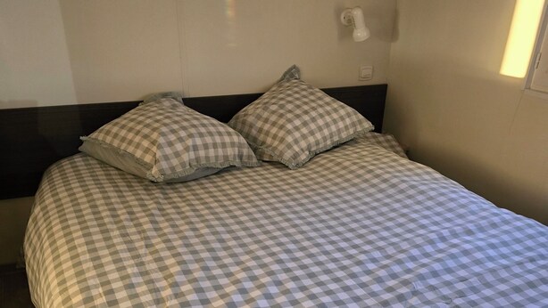 A neatly made bed with a checkered bedspread and two pillows, a bedside table with a lamp, and a wall-mounted light fixture. - Gite in the heart of nature 15 minutes from Colmar  (Widensolen)