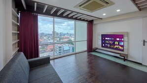 A modern living room with a large window, a flat-screen TV, and a grey sofa. - Spacious 3-bedroom water front apartment in serene Marine Drive Ernakulam Kerala (ERNAKULAM)