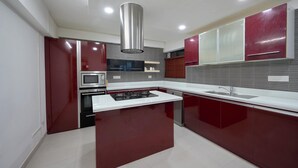 A modern kitchen with red cabinetry, a white countertop, stainless steel appliances, and recessed ceiling lighting. - Spacious 3-bedroom water front apartment in serene Marine Drive Ernakulam Kerala (ERNAKULAM)