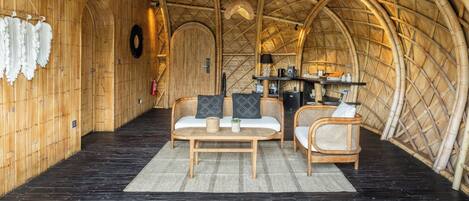 An arched bamboo ceiling, a wooden wall with a hanging white cloth, a round woven rug, and a wooden table with cushions.