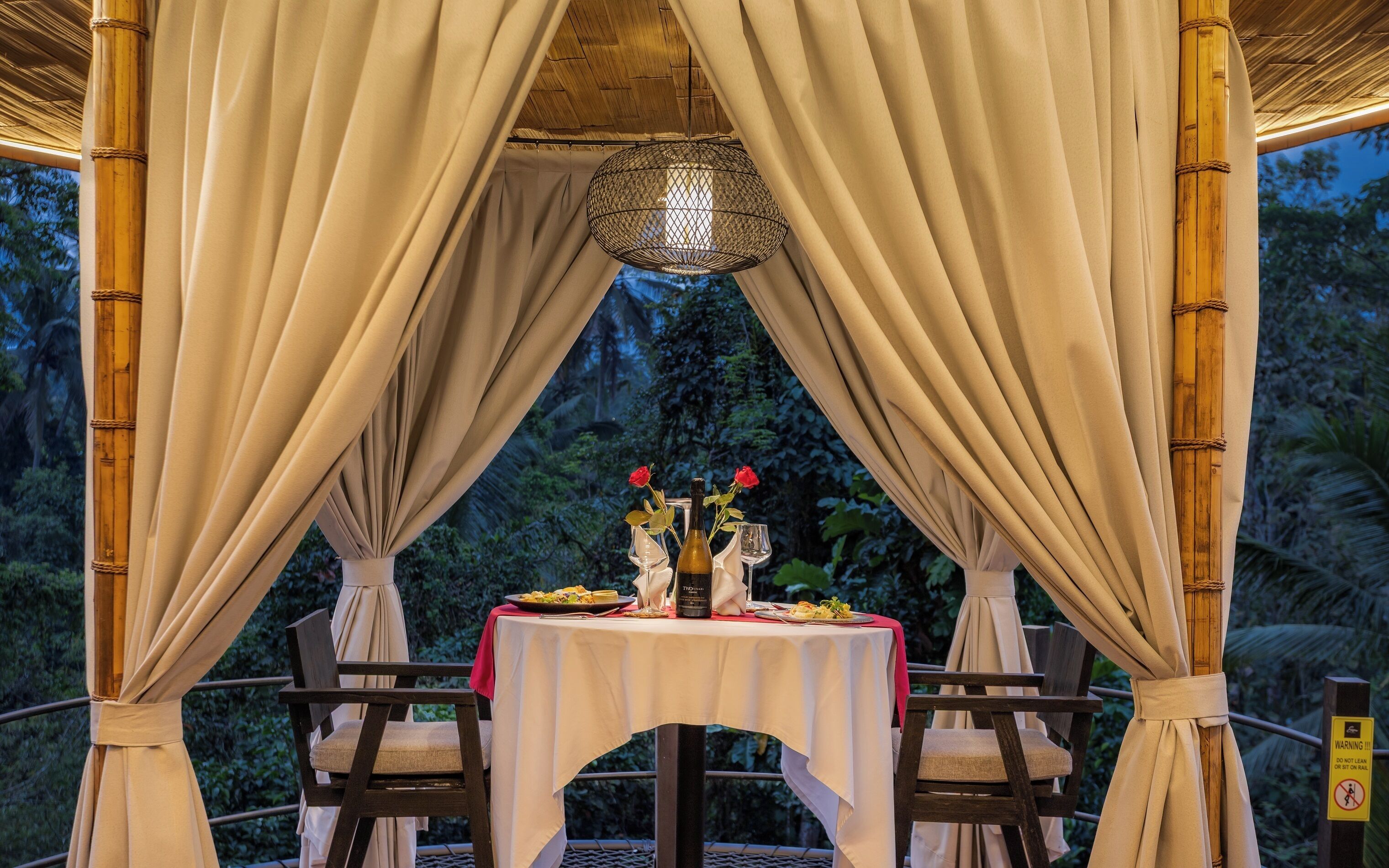 An outdoor dining area with a table set for two, surrounded by bamboo pillars and draped curtains.