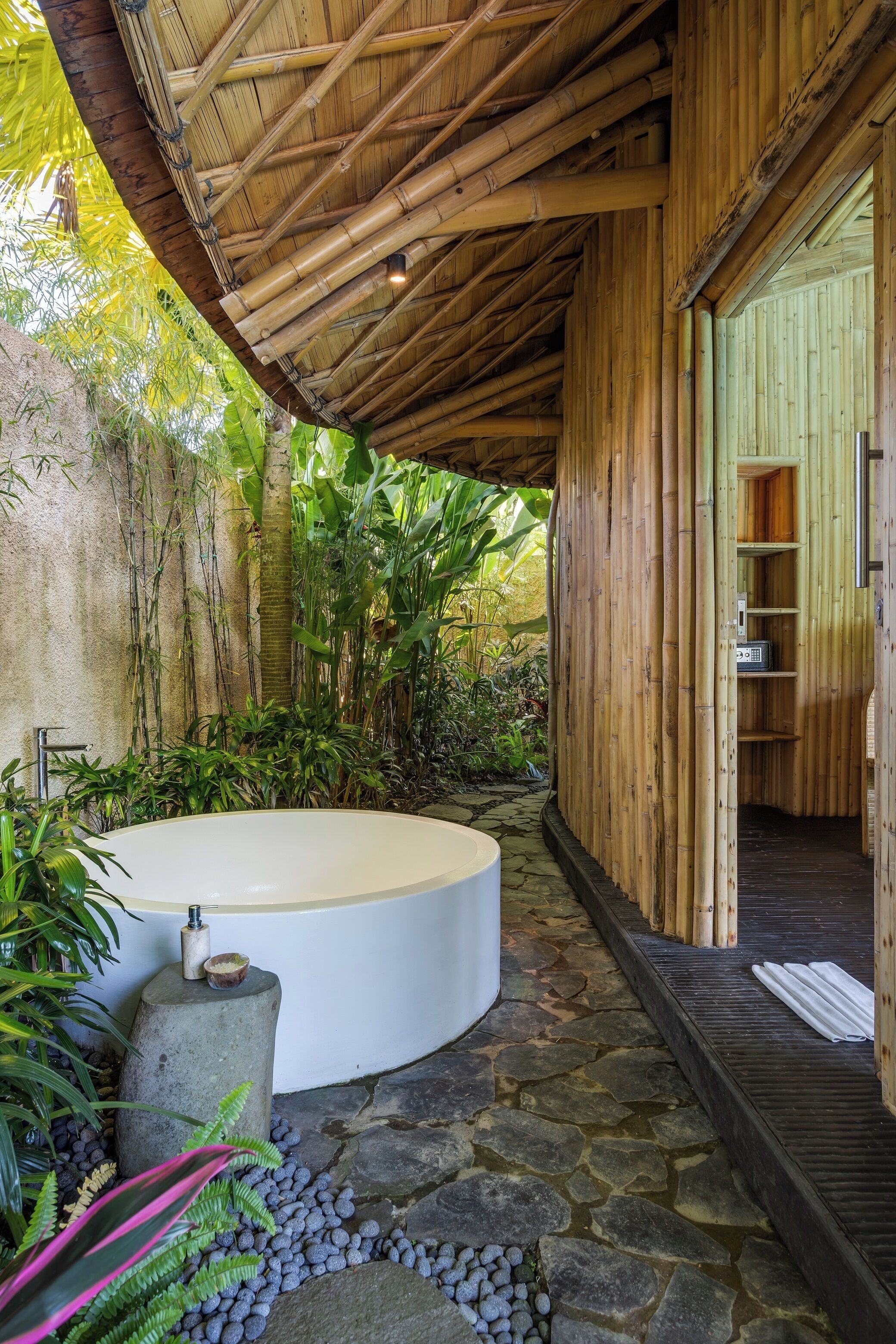 A spa area with a stone pathway leading to a circular hot tub, surrounded by lush greenery and bamboo structures.