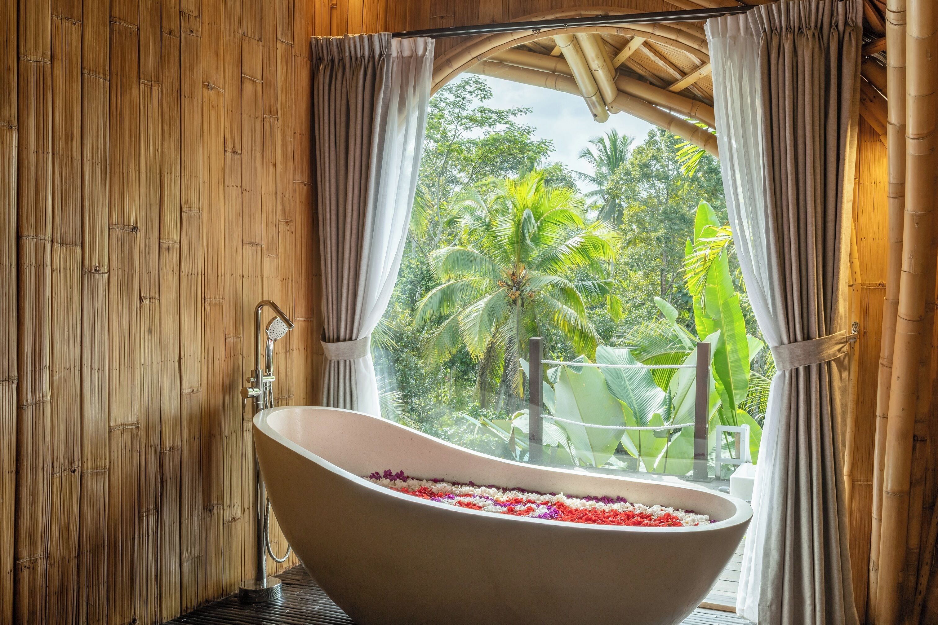 A bathroom with a freestanding bathtub filled with flower petals, a bamboo wall, and a view of lush greenery outside.