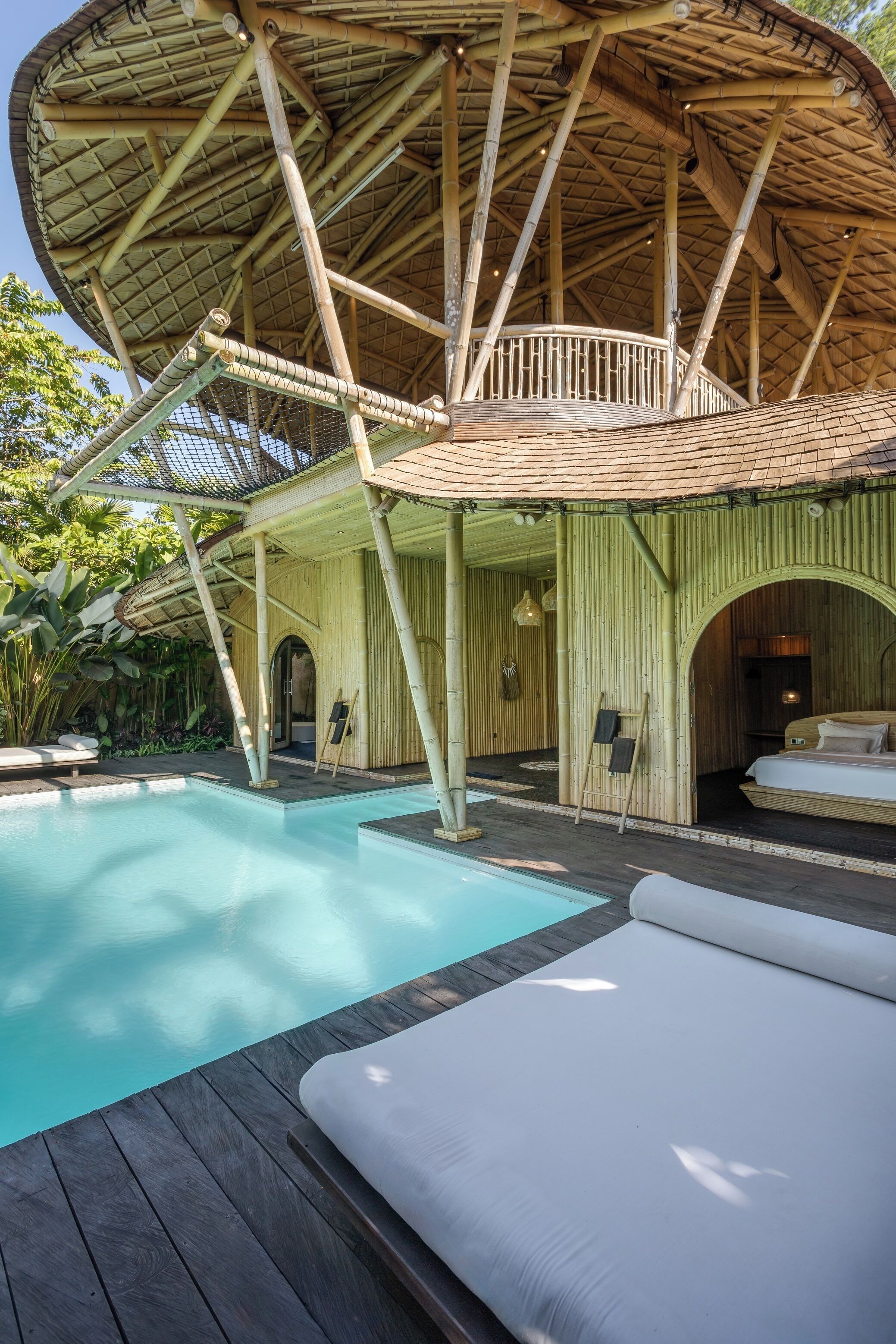 A pool area with a wooden deck and lounge chairs, a bamboo structure with a thatched roof, and a bed with white bedding.