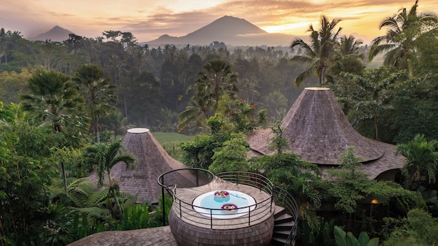 A circular wooden structure with a central table and a railing, surrounded by lush greenery and palm trees, with a mountain in the background.
