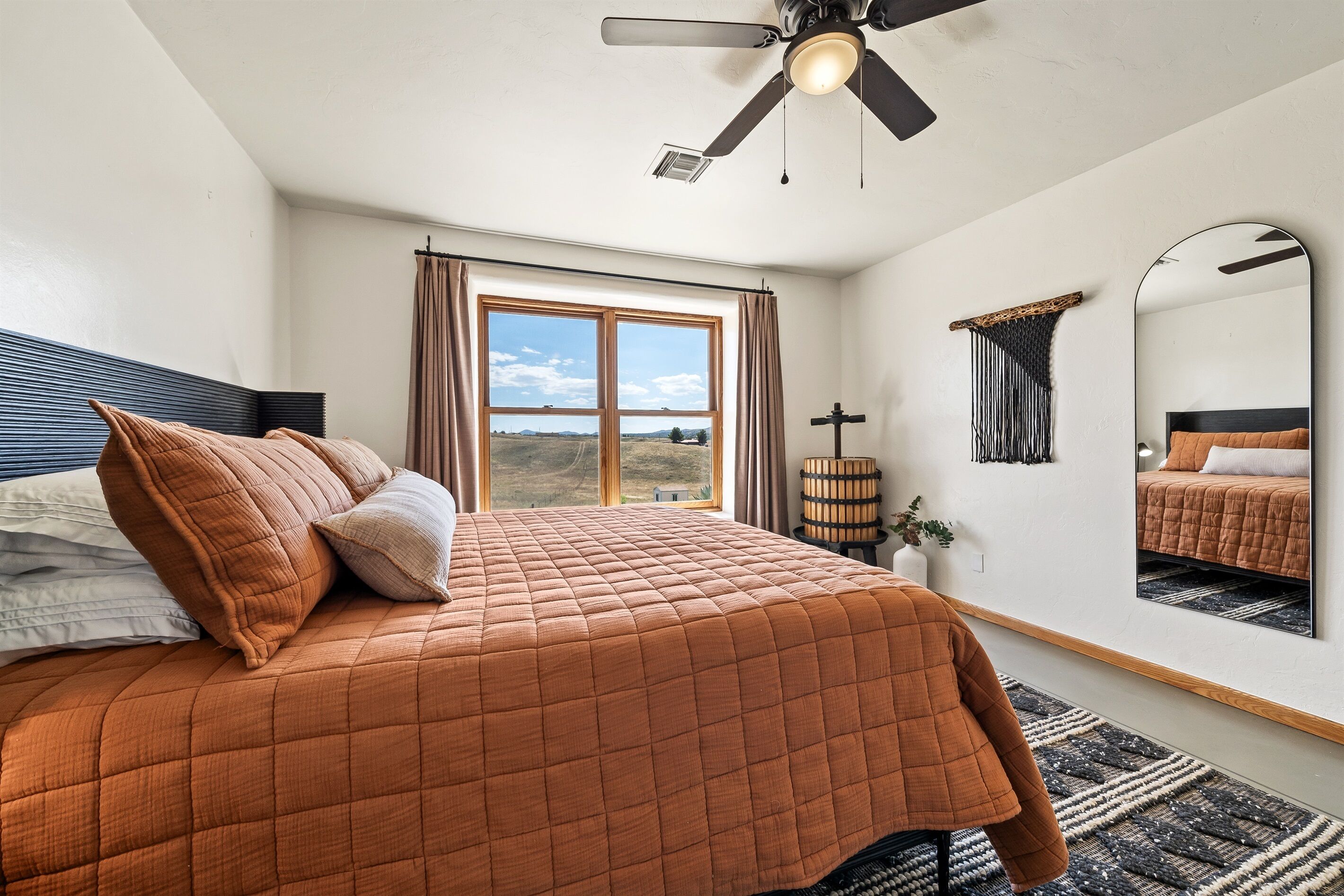 Une chambre dotée d’un grand lit, d’un ventilateur de plafond, d’une fenêtre avec des rideaux, d’un miroir fixé au mur et offrant une vue sur l’extérieur.