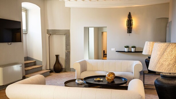 Een moderne woonkamer met een bank, salontafel en decoratieve objecten. Er is een open haard, een aan de muur bevestigde televisie en een trap die naar een hoger niveau leidt.