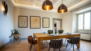 Een eethoek met een houten tafel, vier stoelen en hanglampen. Op de muur hangen kunstwerken in lijsten, er staat een klein bijzettafeltje met een plant en een raam zorgt voor natuurlijk licht.