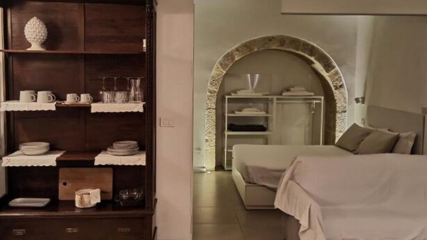 Een slaapkamer met een bed, een houten kast met servies en een stenen boog die naar een badkamer leidt.