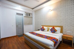 A neatly arranged bedroom with a bed, a nightstand, a wooden floor, a white brick wall, and a door. - Fabhotel Prime Culture (Noida)