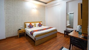 A hotel room with a brick-patterned wall, a wooden bed with colorful pillows, a small bedside table, a desk with a chair, and a ceiling fan. - Fabhotel Prime Culture (Noida)