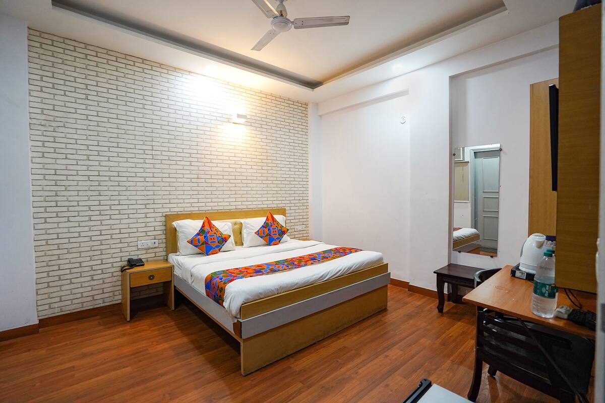Habitación de hotel con pared de ladrillo, cama de madera con almohadas de colores, mesita de noche pequeña, escritorio con silla y ventilador de techo.