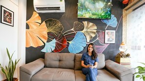 Un salon moderne avec un canapé gris, une petite table basse en bois et une grande fresque murale représentant des feuilles stylisées.
