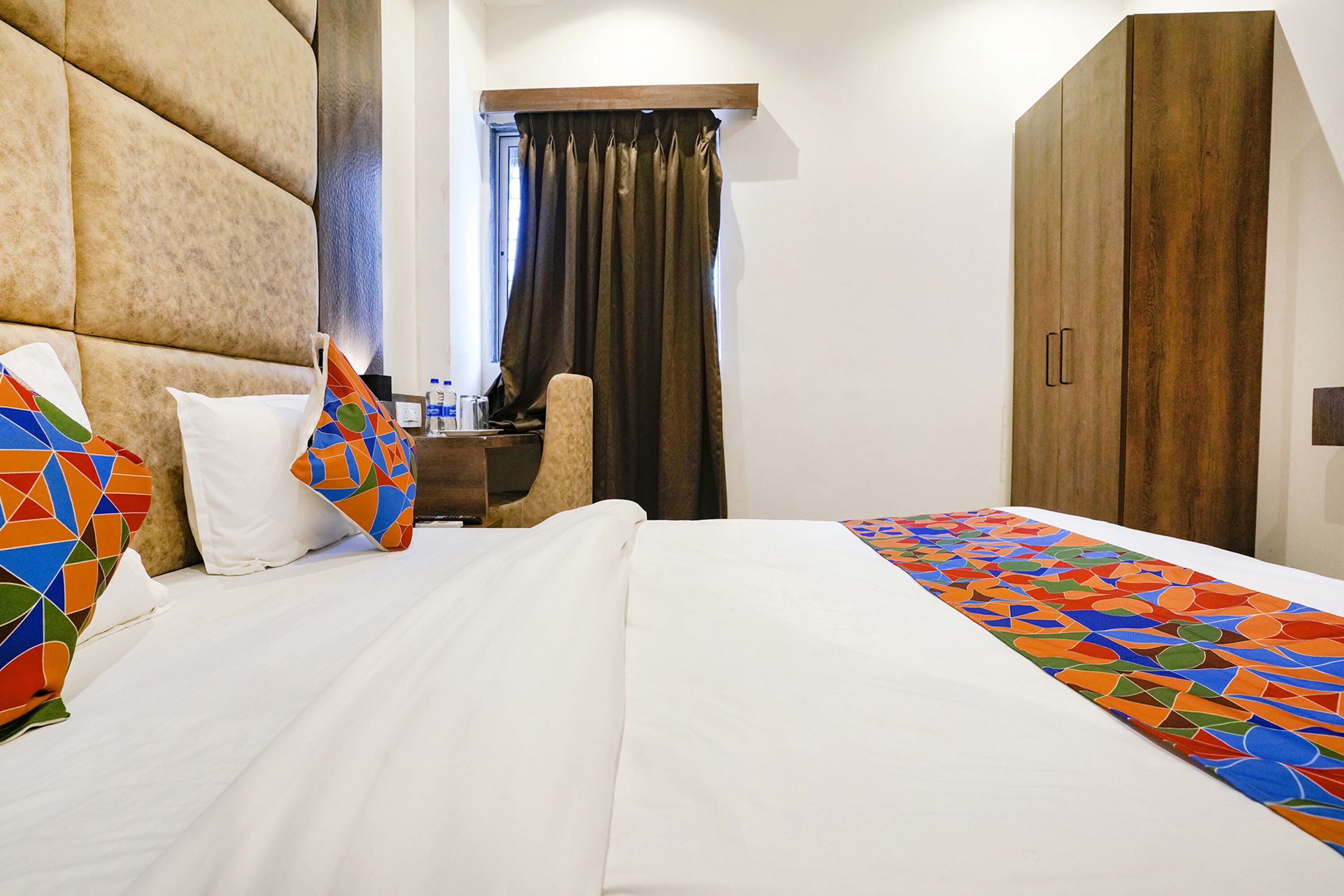 A hotel room with a large bed, a wooden wardrobe, a desk with a chair, and a window with curtains.