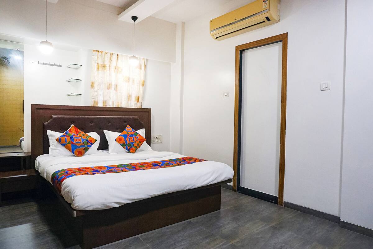 A neatly made bed with a wooden headboard, a colorful bedspread, and decorative pillows. There is a bedside table with a lamp, and a wall-mounted air conditioner.