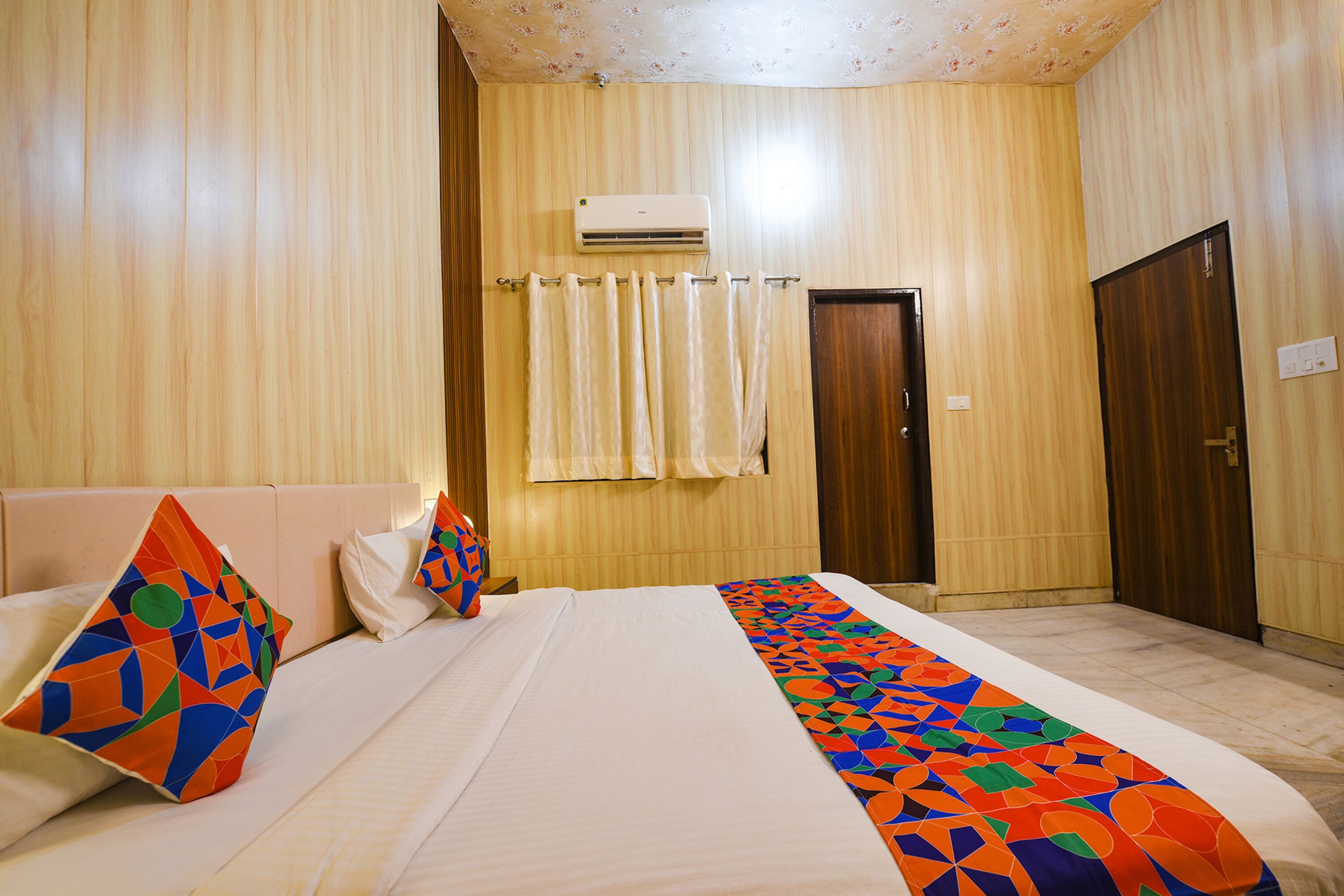 Una habitación de hotel con cama, almohadas, colcha estampada, equipo de aire acondicionado, una puerta y una cortina.