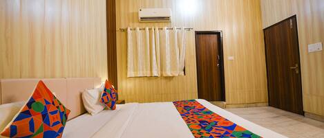 Una habitación de hotel con cama, almohadas, colcha estampada, equipo de aire acondicionado, una puerta y una cortina.