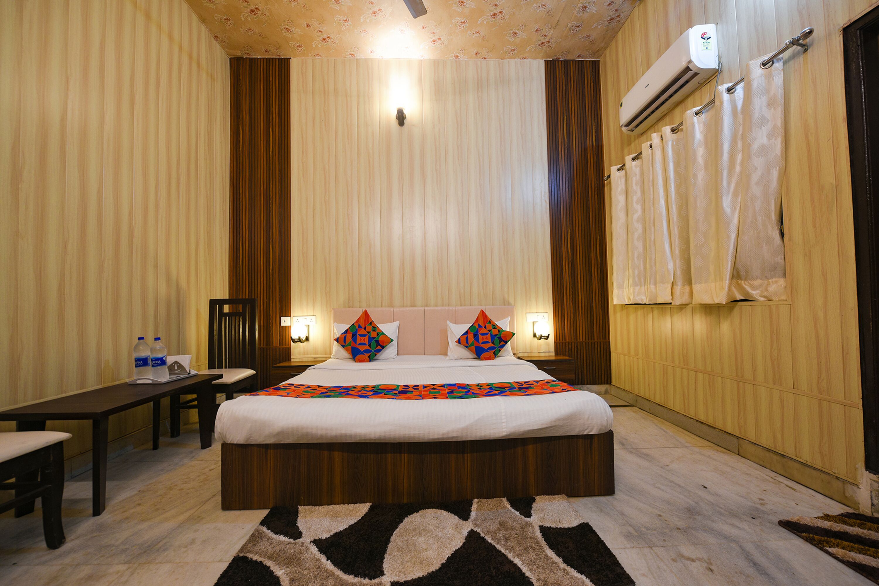 Une chambre d’hôtel avec un lit, des tables de chevet, un bureau et une chaise. Il y a un climatiseur mural et une fenêtre avec des rideaux.