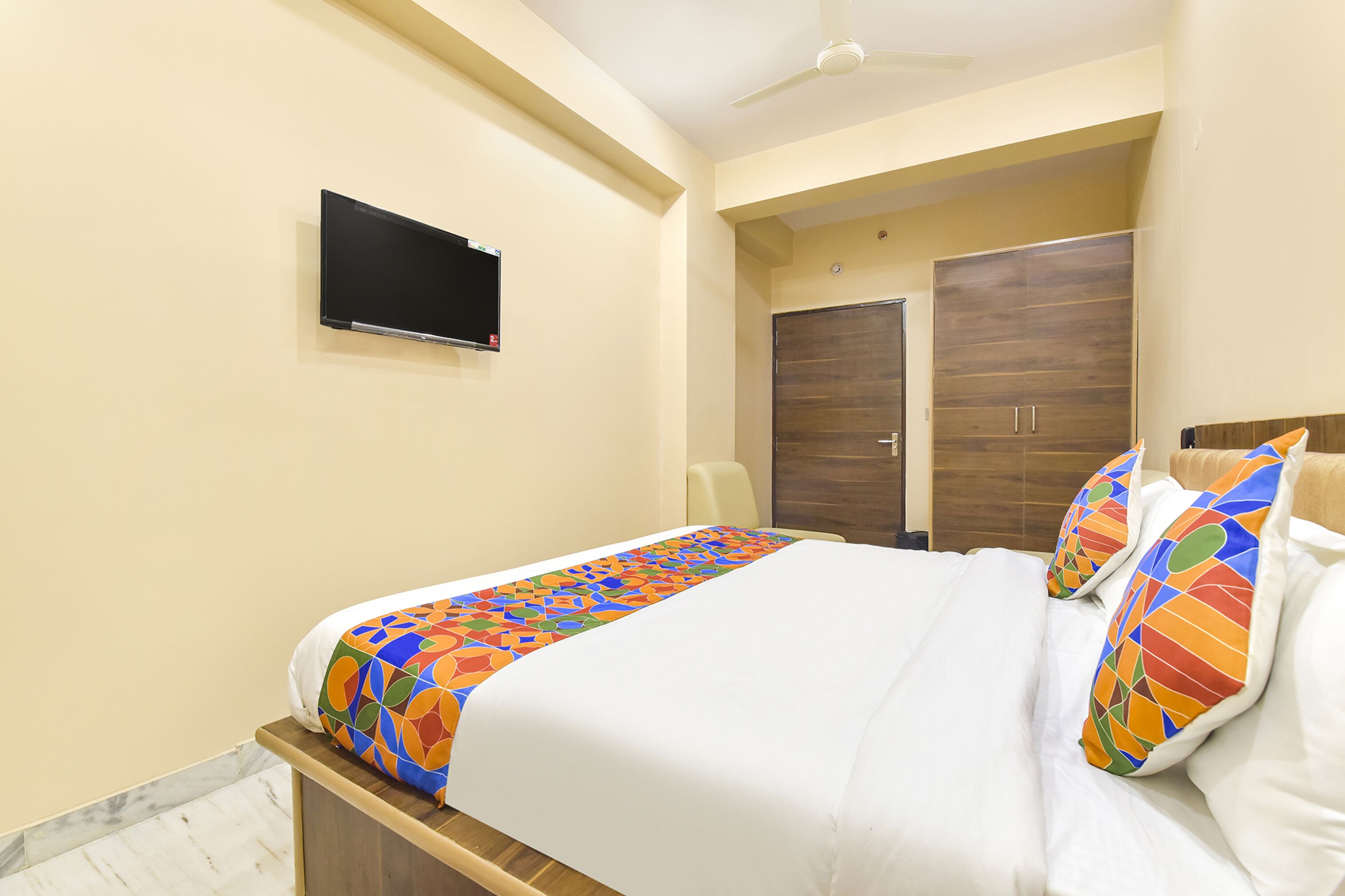A neatly made bed with a colorful geometric-patterned bedspread and matching pillows, a flat-screen TV mounted on the wall, and a wooden wardrobe with a mirror.