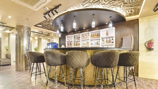 A bar area with a round counter, four cushioned stools, and a shelf with various bottles. - Fabhotel Pearl Square (Jaipur)