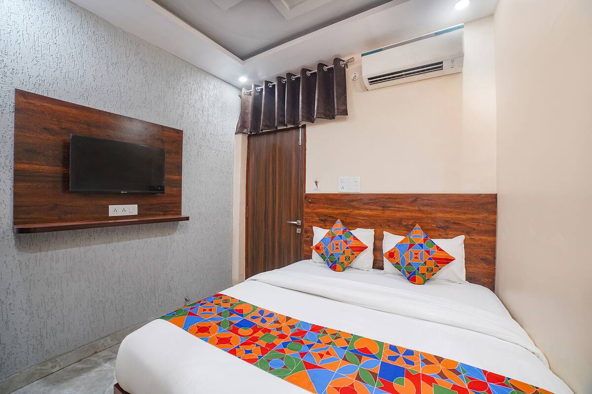 Una habitación de hotel con cama, televisor montado, cabecera de madera y colcha estampada.