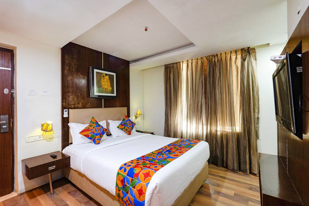 A hotel room with a bed, a nightstand, a TV, a painting on the wall, and a window with curtains.