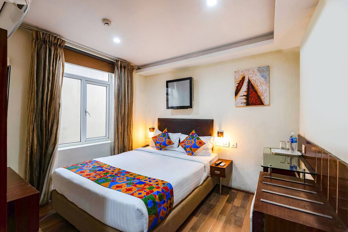 A hotel room with a bed, a nightstand, a painting on the wall, and a window with curtains.