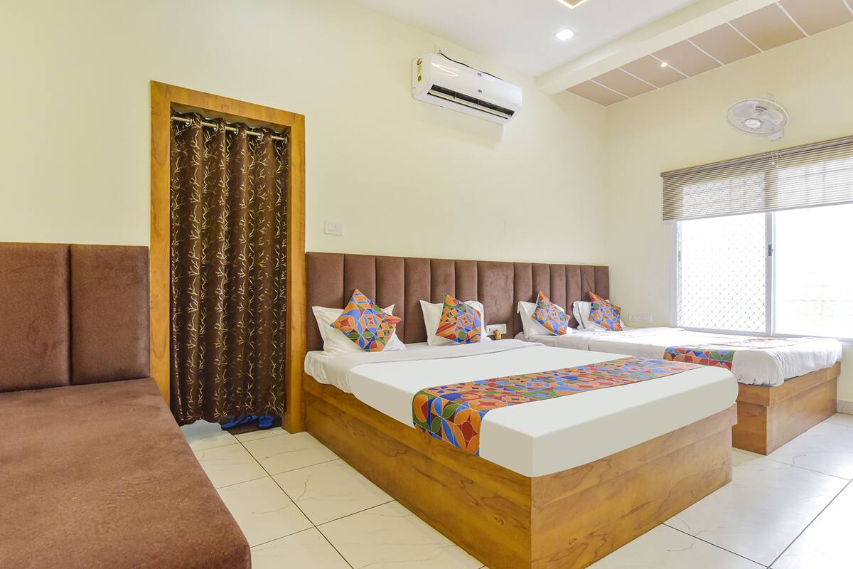 A hotel room with a large bed, a smaller bed, a sofa, an air conditioning unit, a ceiling fan, and a window with blinds.