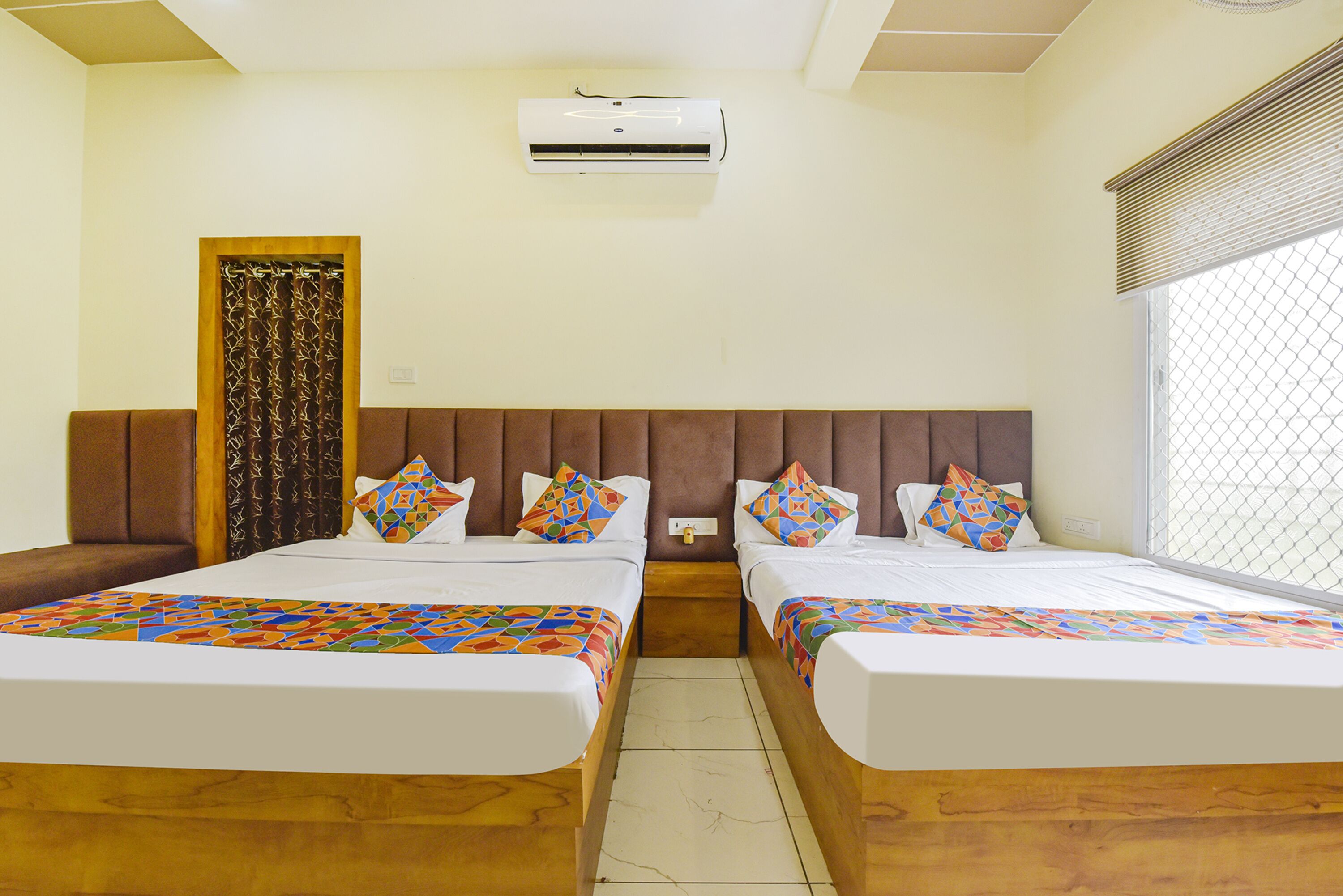Habitación de hotel con dos camas, cabecero de madera, unidad de aire acondicionado, ventana con persianas y cortina decorativa.