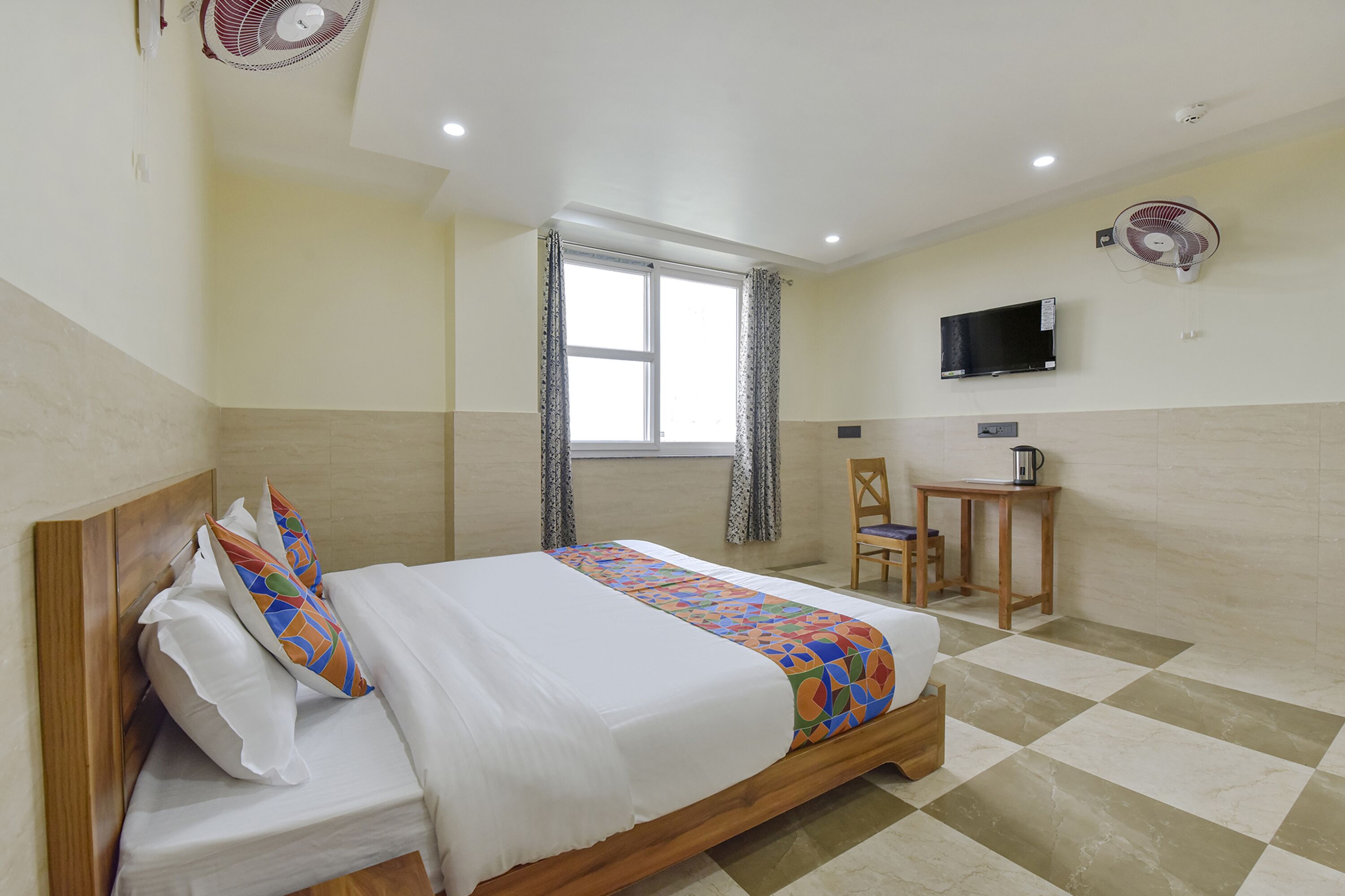 A hotel room with a bed, a desk, a chair, a TV, and two wall fans.