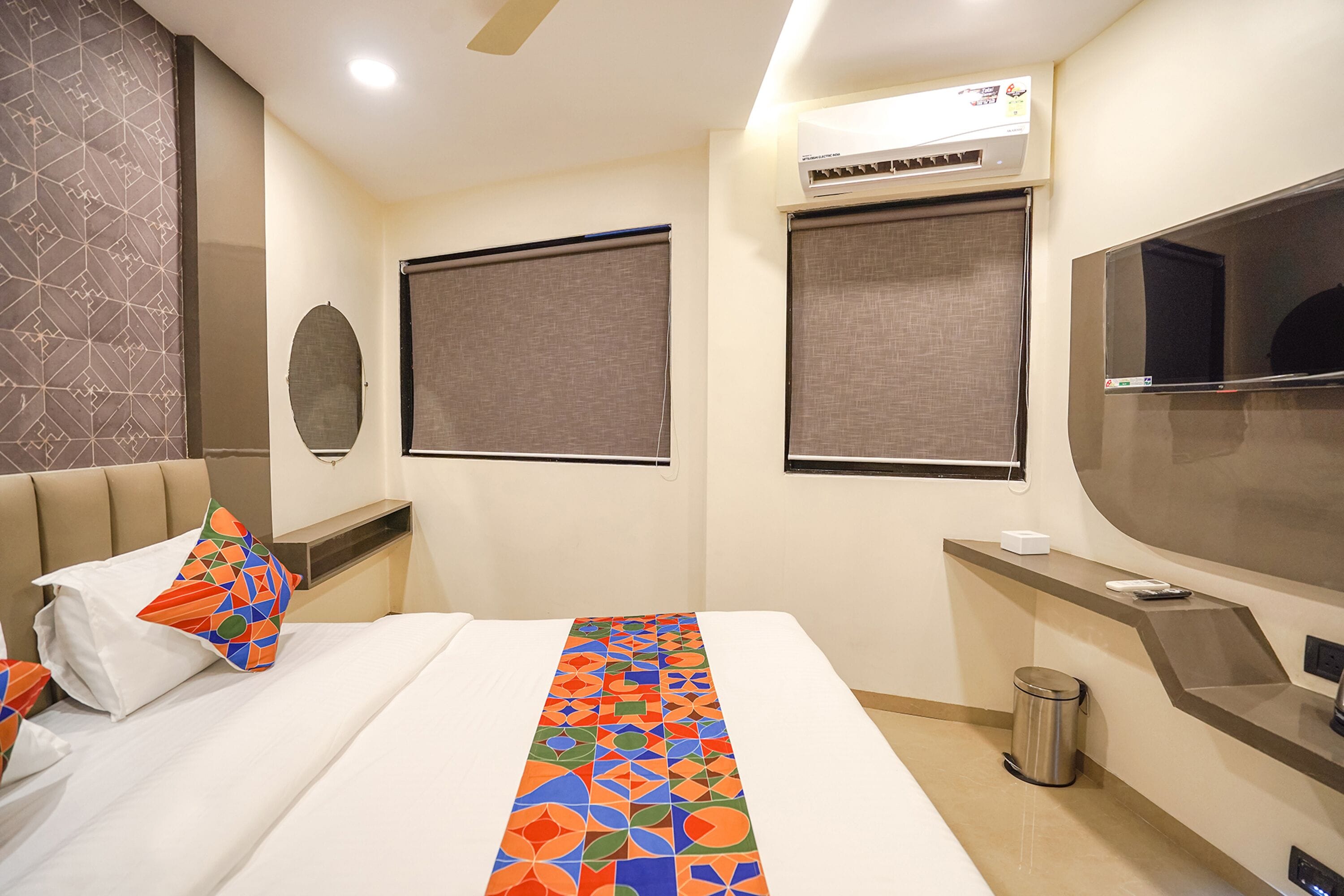 A hotel room with a bed, a TV, an air conditioning unit, and a trash bin.