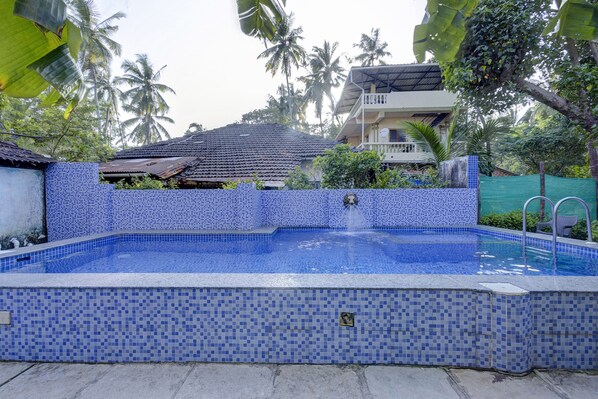 A swimming pool with blue mosaic tiles, surrounded by greenery and a building with a tiled roof. - Fabhotel Arya'S Hyderabad Resort (Calangute)