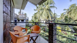 Un balcón con dos sillas naranjas y una mesa pequeña, con vista a un jardín exuberante con palmeras y edificios al fondo.