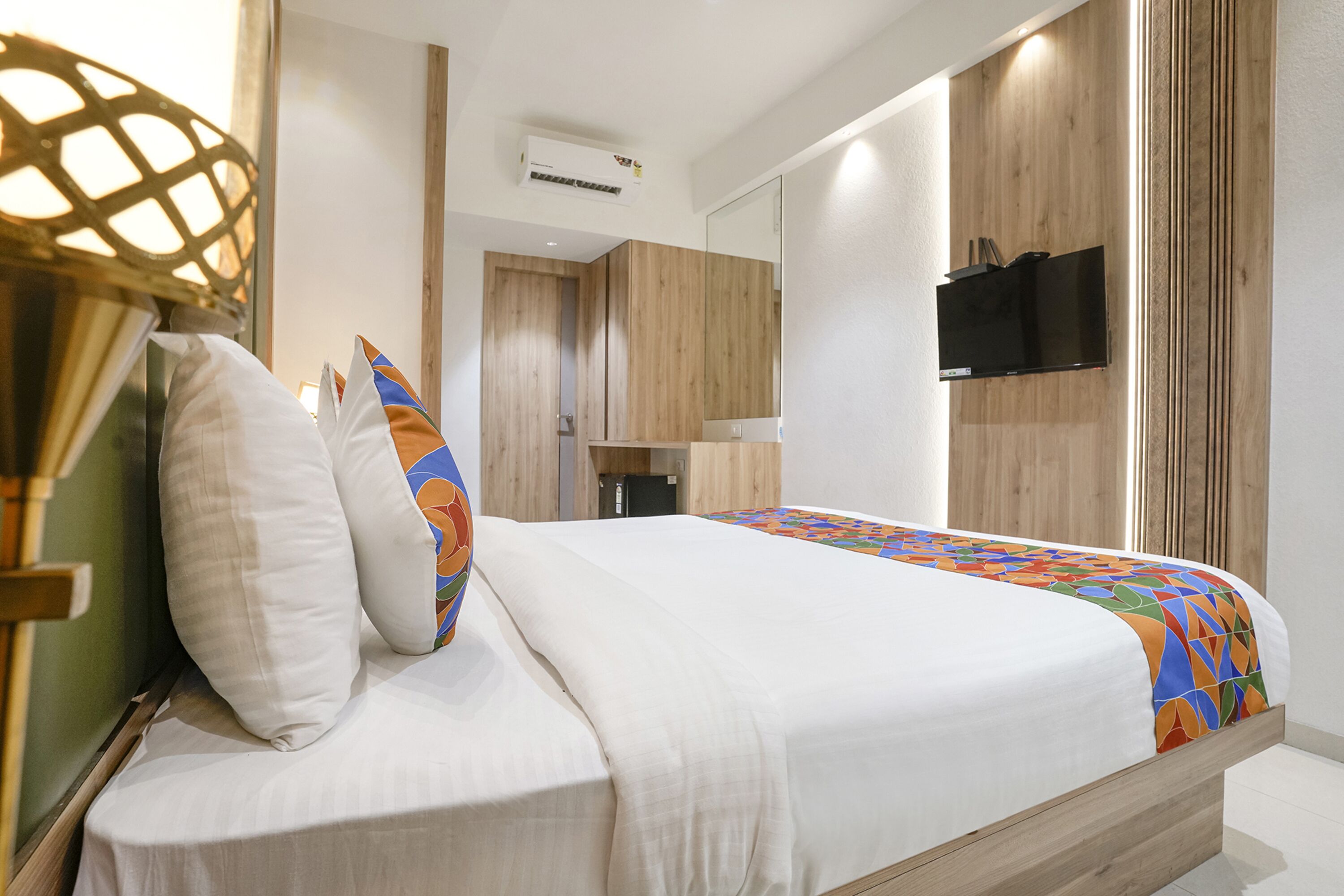 Une chambre à coucher moderne avec un cadre de lit en bois, un matelas blanc et une courtepointe aux couleurs vives. Un téléviseur est fixé au mur, et une petite table de chevet avec une lampe se trouve à côté du lit.