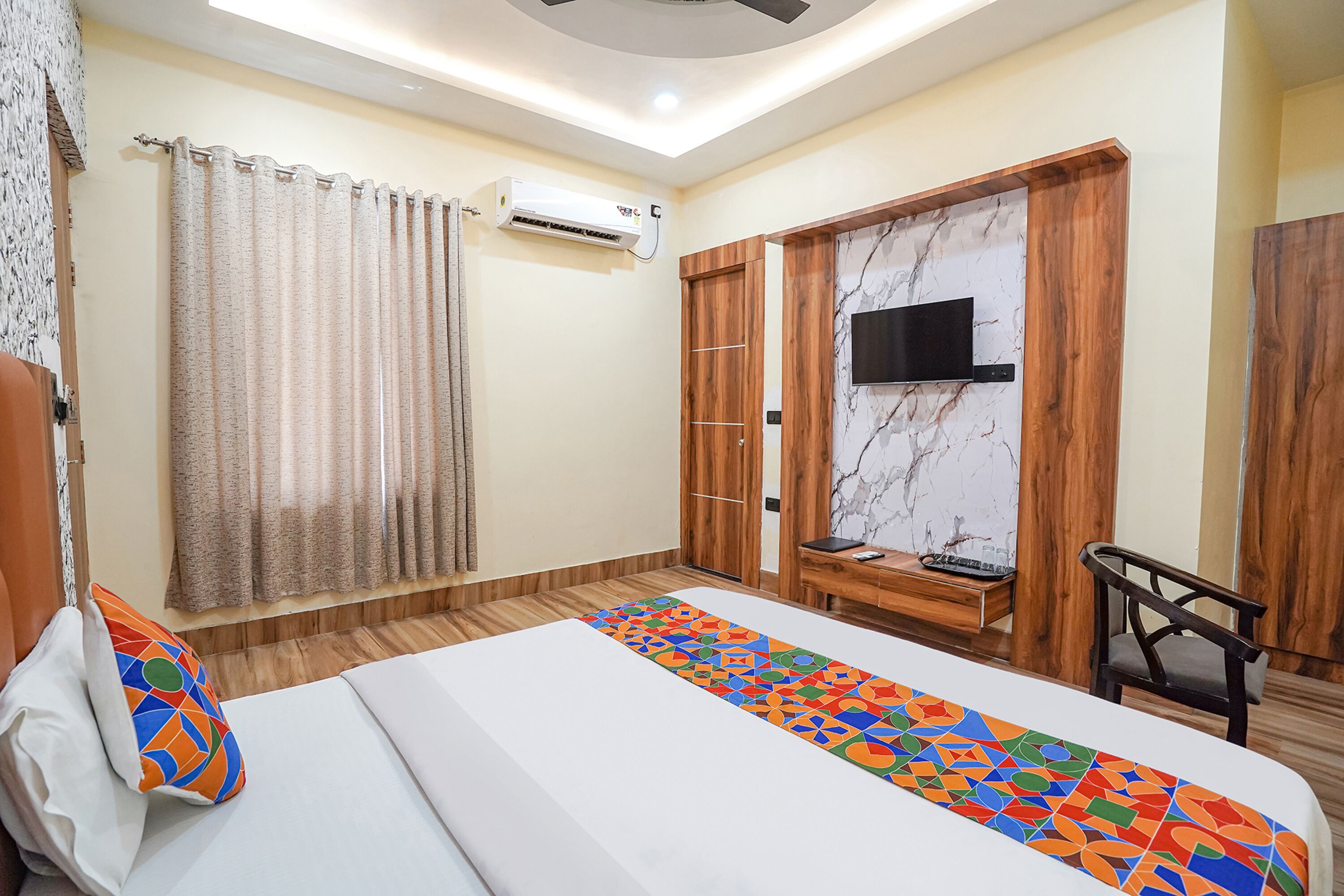 A hotel room with a bed, a TV mounted on a wooden wall, an air conditioning unit, and a desk with a chair.