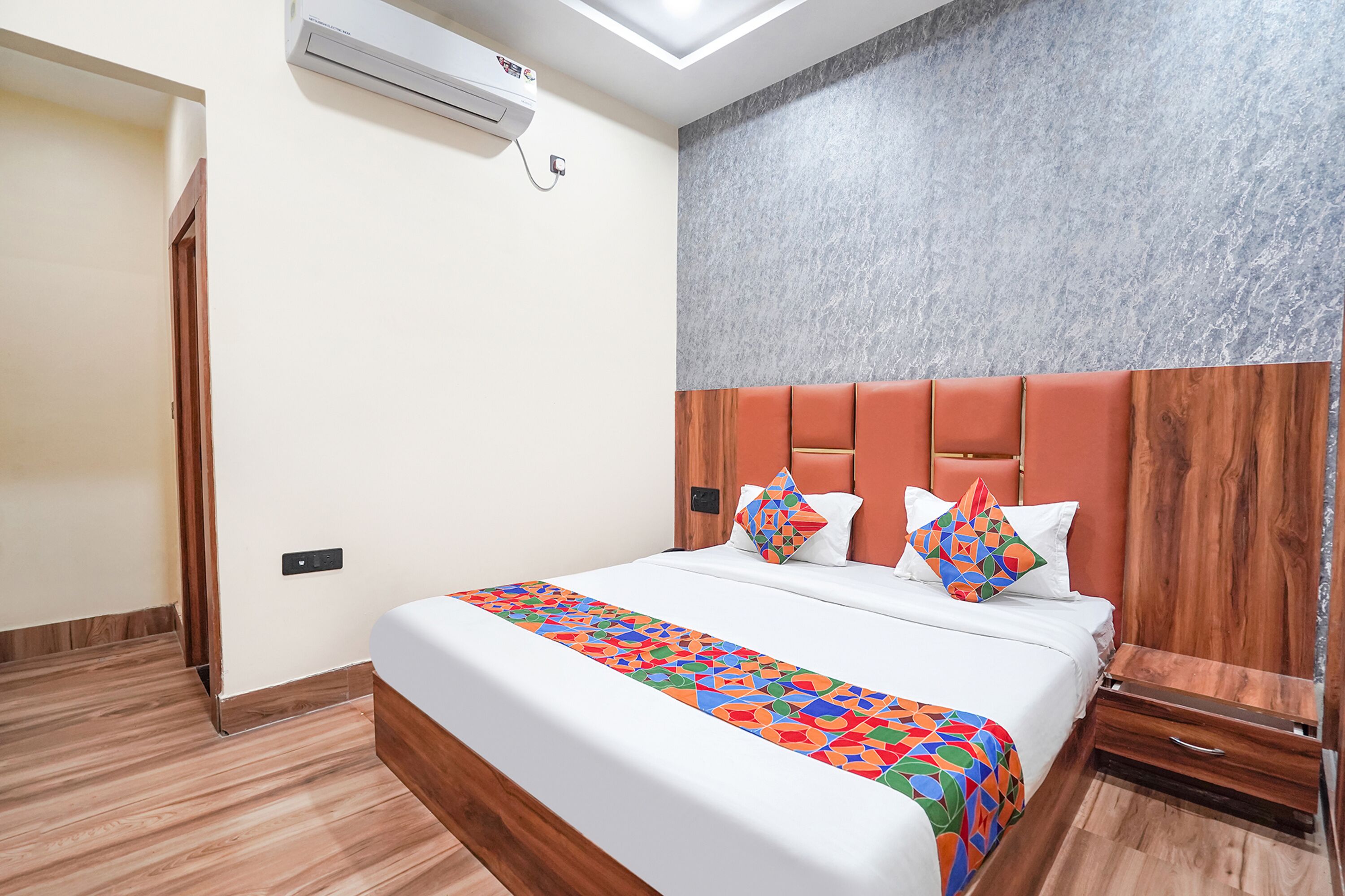 A modern hotel room with a bed, a wooden headboard, a nightstand, and an air conditioning unit.