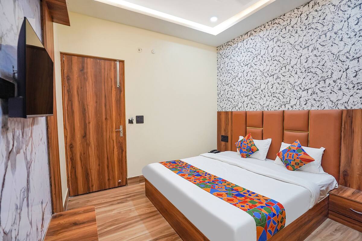 A hotel room with a wooden door, a bed with a colorful geometric bedspread, a headboard with a patterned wallpaper, and a built-in TV on the wall.