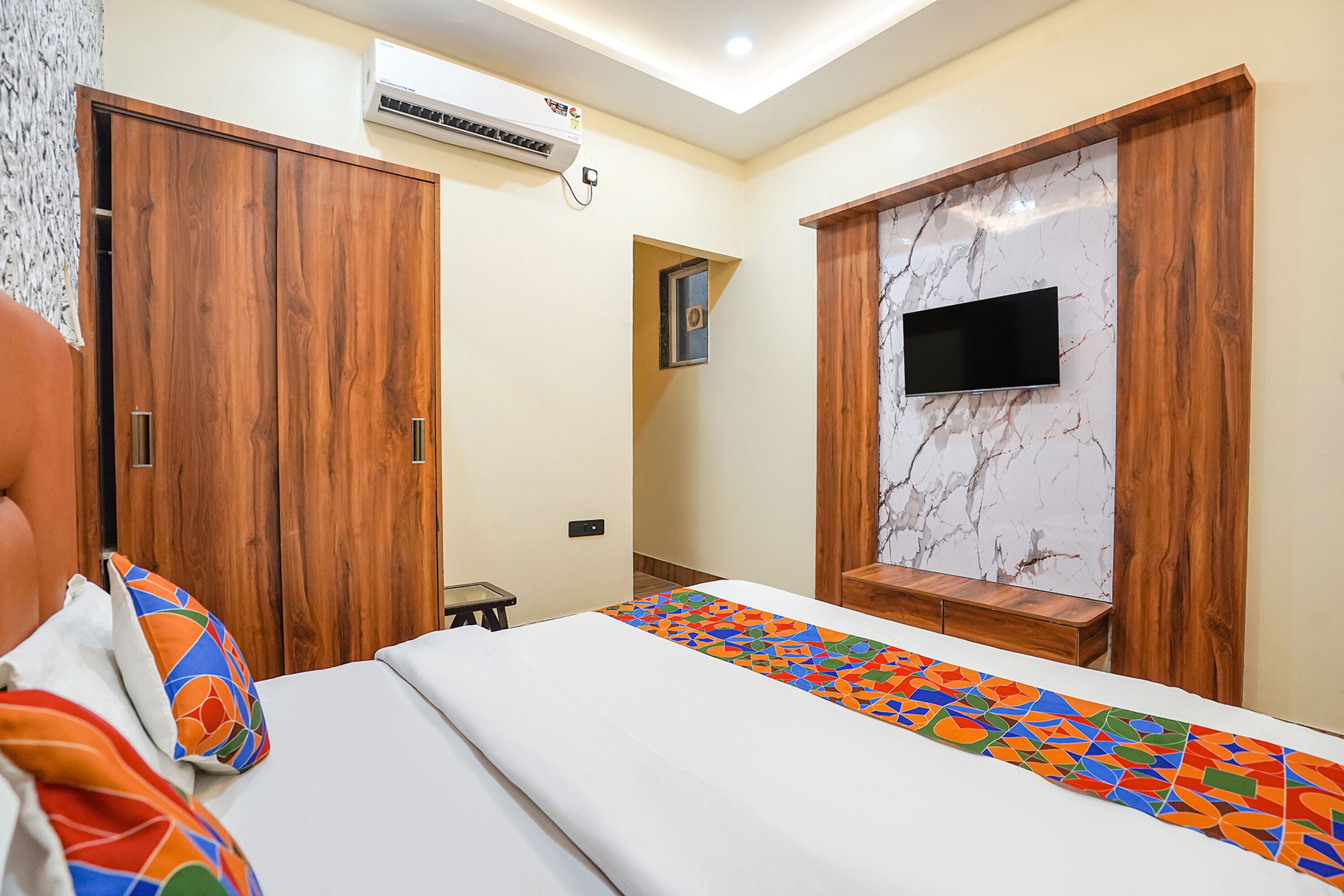 A bedroom with a bed, a TV mounted on a marble wall, wooden wardrobes, and an air conditioning unit.
