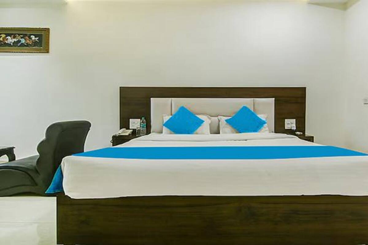 A neatly made bed with a blue bedspread and two blue pillows, a wooden headboard, a nightstand with a phone and a bottle, and a chair to the left.