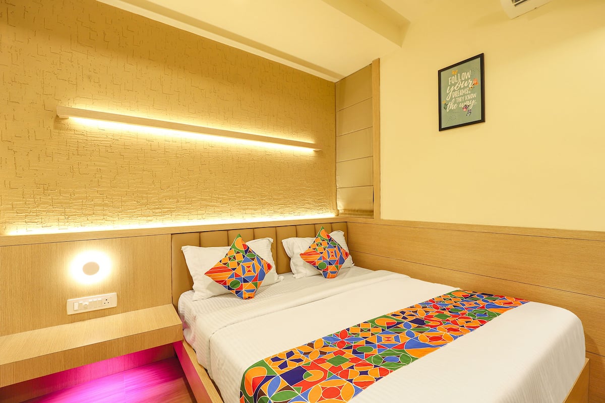 A modern hotel room with a wooden headboard, a bed with white linens, colorful geometric-patterned pillows, and a wall-mounted light fixture.