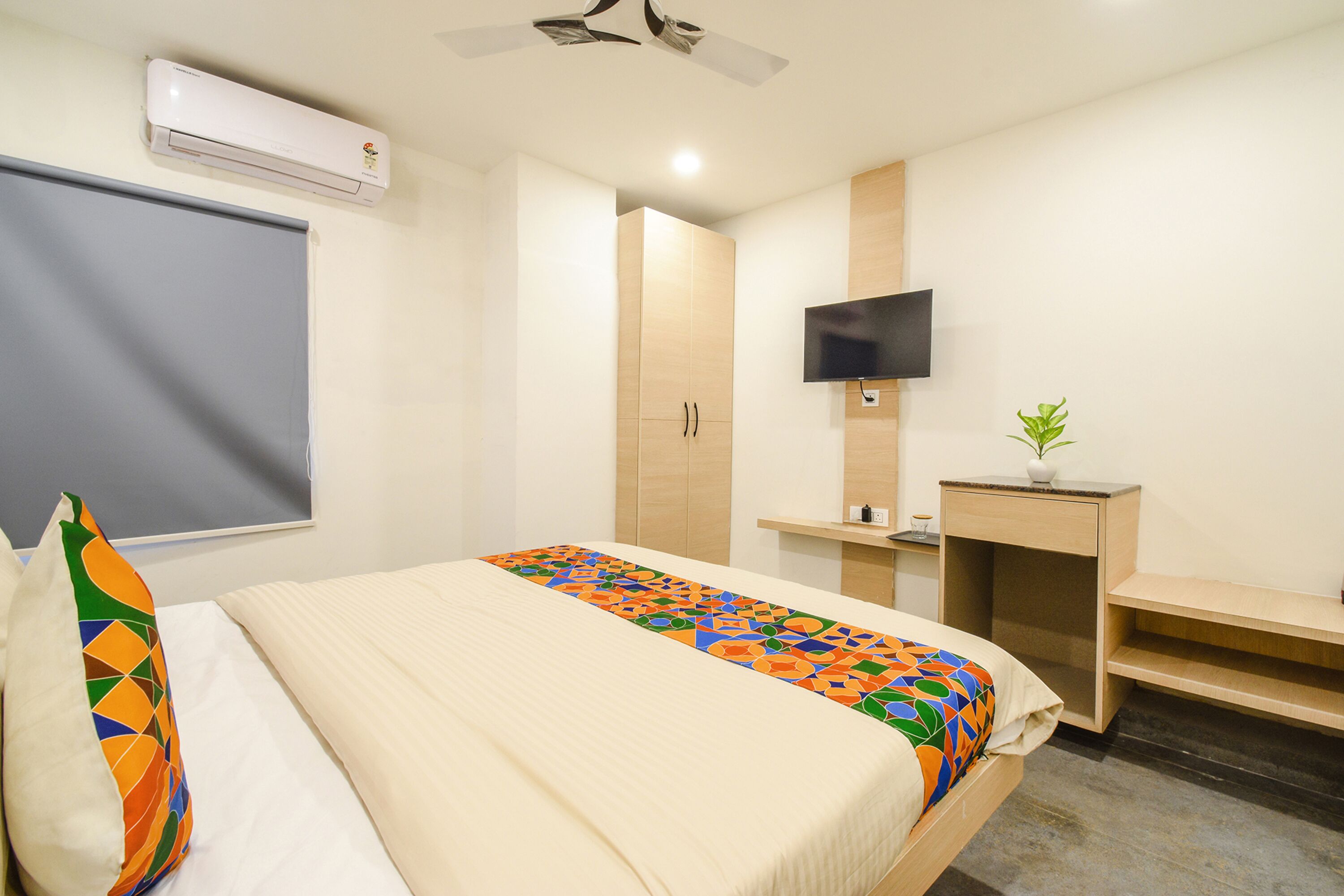 A hotel room with a bed, a TV mounted on the wall, a desk with a plant, and an air conditioning unit.