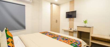 A hotel room with a bed, a TV mounted on the wall, a desk with a plant, and an air conditioning unit.