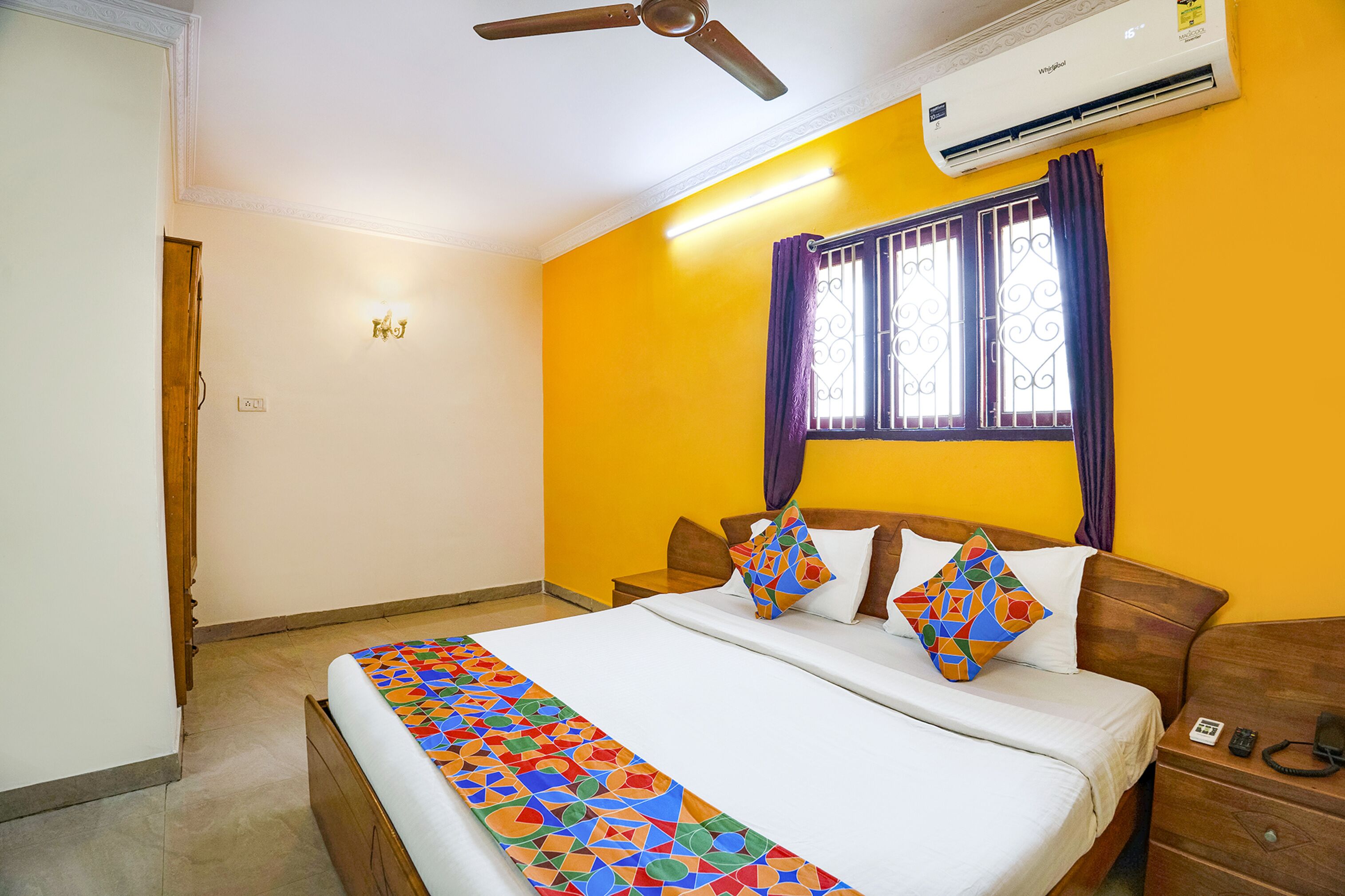 A hotel room with a bed, a nightstand, a ceiling fan, an air conditioning unit, and a window with decorative bars and curtains.