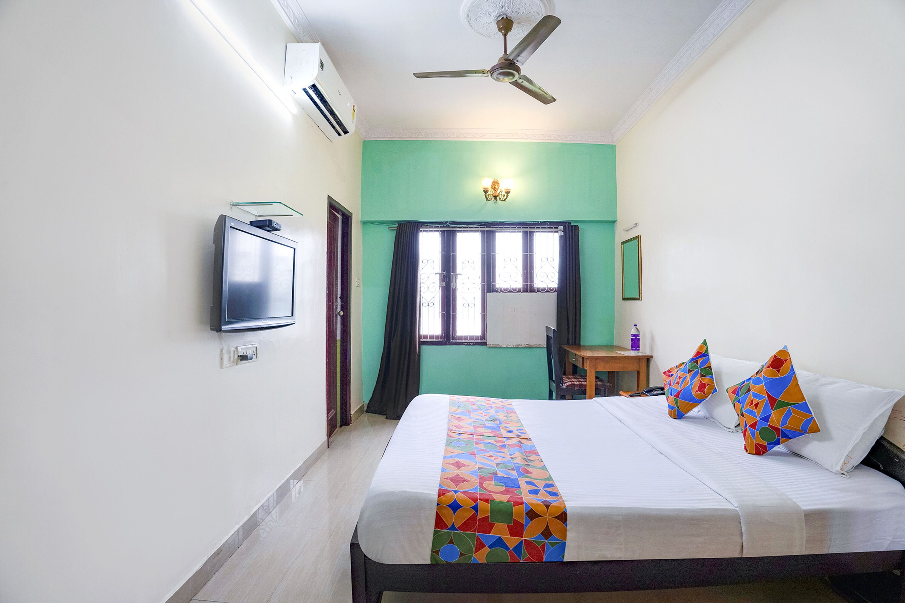 Une chambre d’hôtel avec un lit, un ventilateur de plafond, une télévision, une fenêtre avec des rideaux et une petite table avec une chaise.