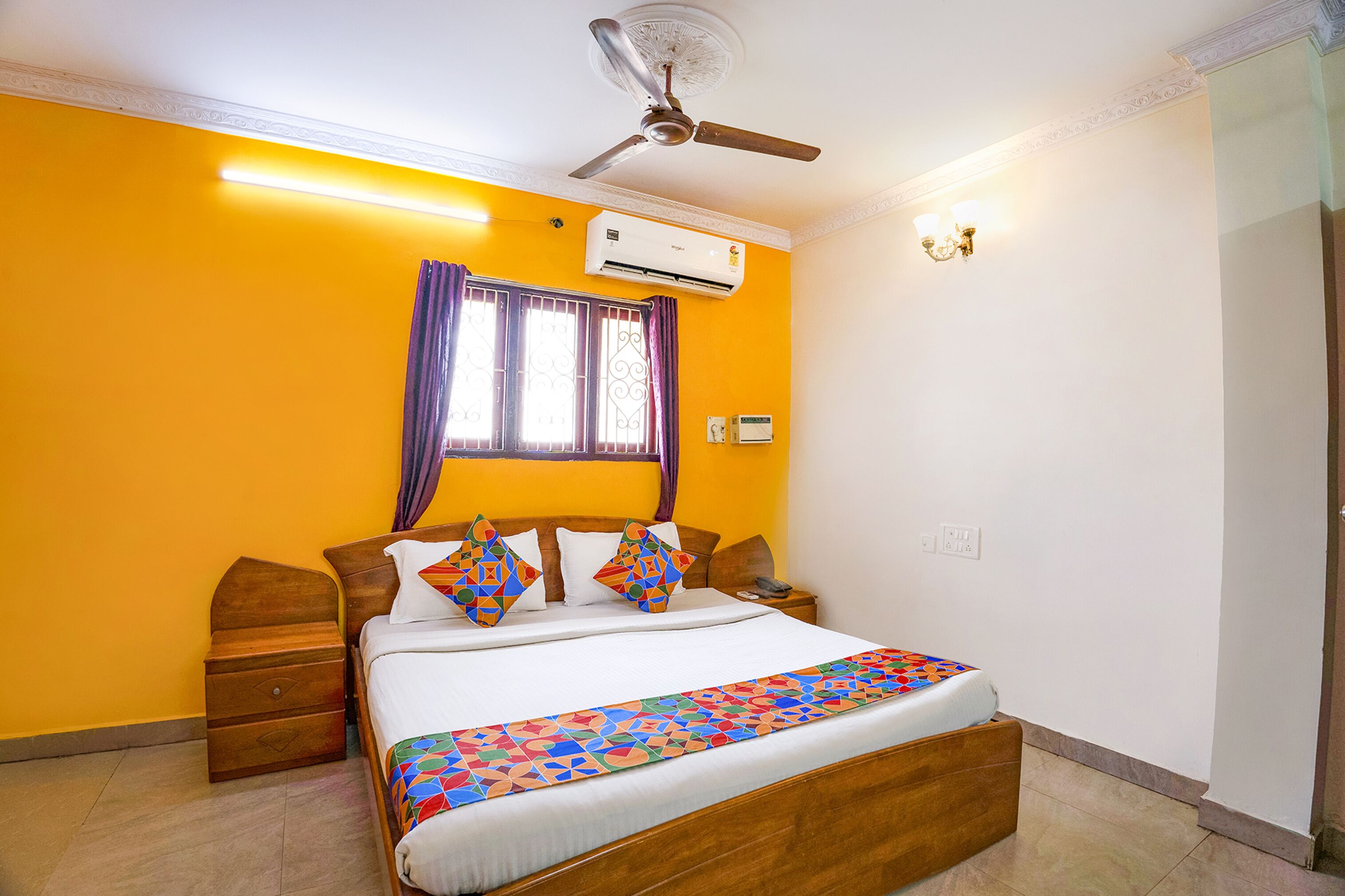 A bedroom with a bed, a ceiling fan, an air conditioning unit, a nightstand, and a window with purple curtains.