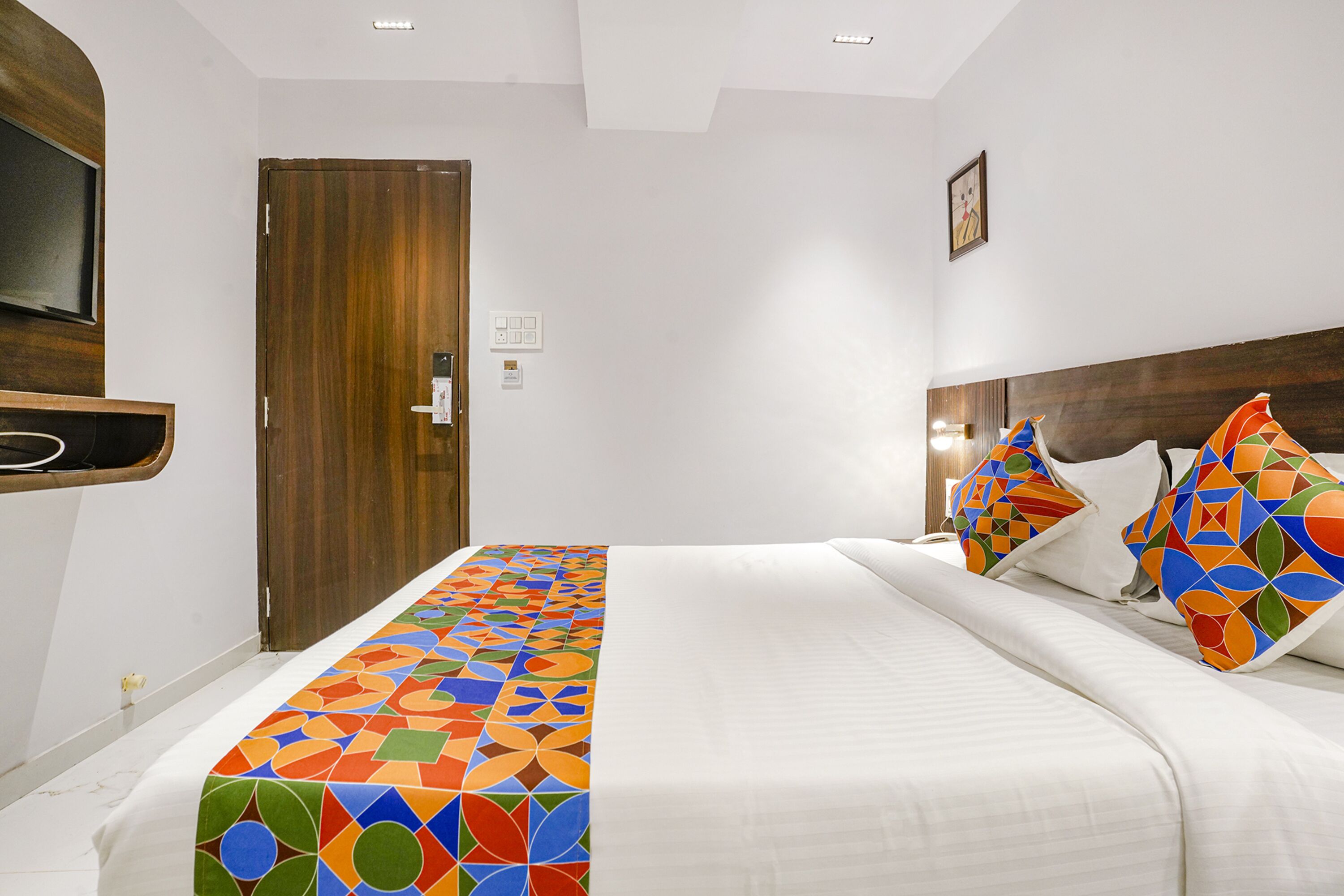 A hotel room with a bed, a wooden door, a TV mounted on the wall, and a colorful bedspread.